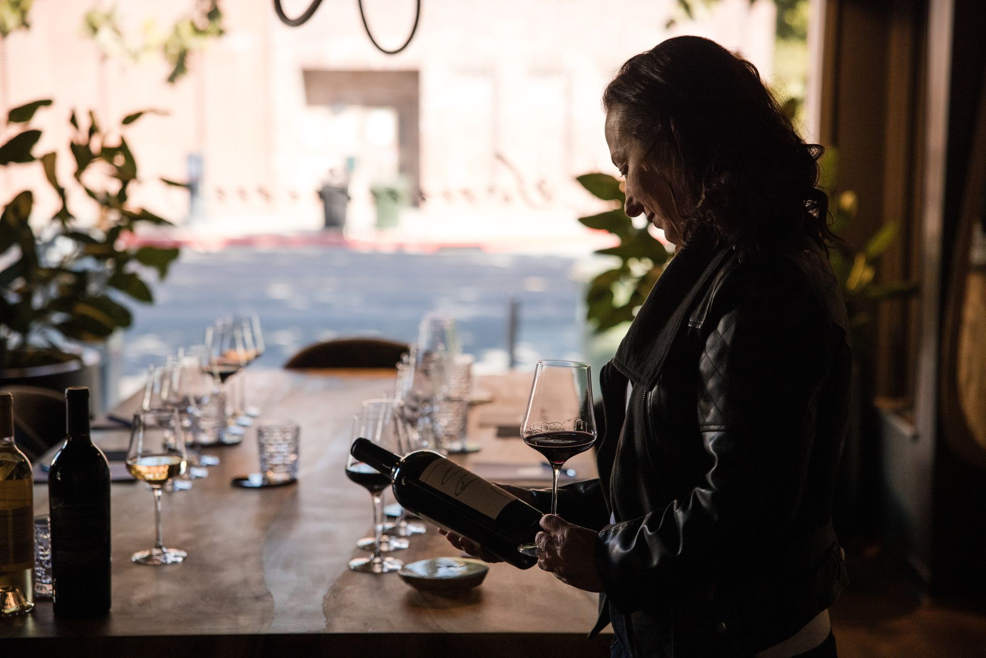 Woman in leather jacket examining a wine bottle at a table with glasses in a wine tasting room.
