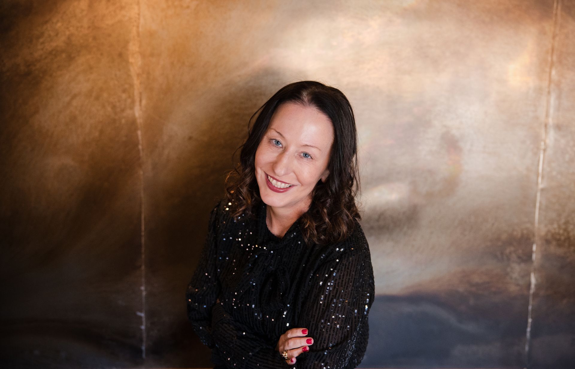 Woman with dark hair smiles, wearing a black sequined top, posing in front of a textured gold wall.