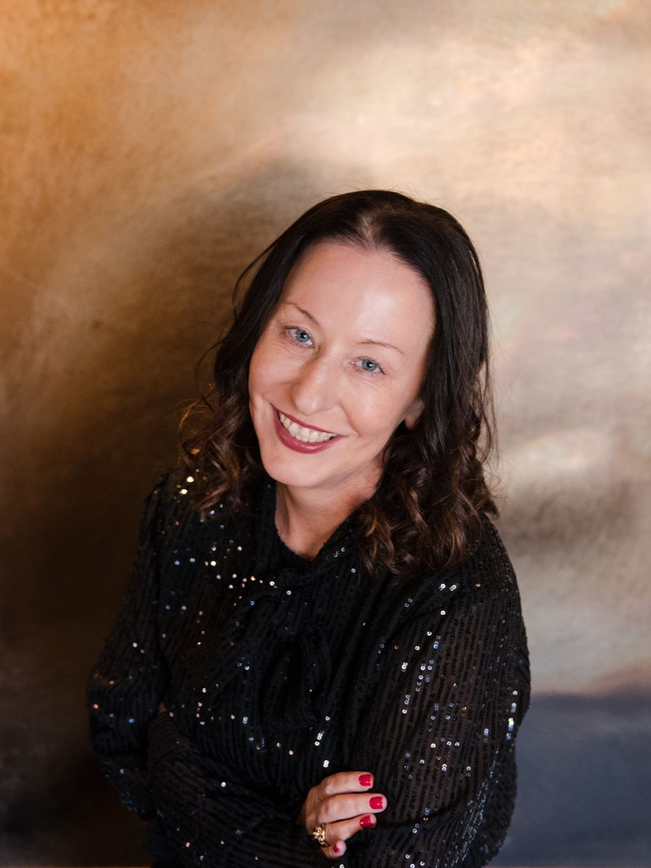 Woman with dark hair smiles, wearing a black sequined top, against a mottled brown background.