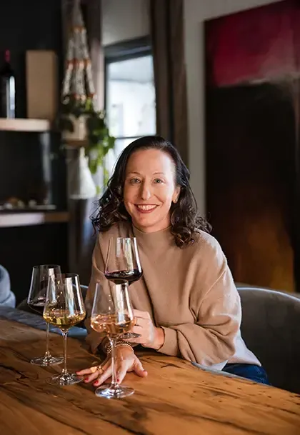 Woman at wooden table holding wine glass, smiles. Three wine glasses, various wines. Interior setting.