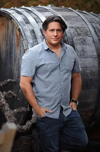 Man in blue shirt and jeans leans against a large wooden barrel outdoors.