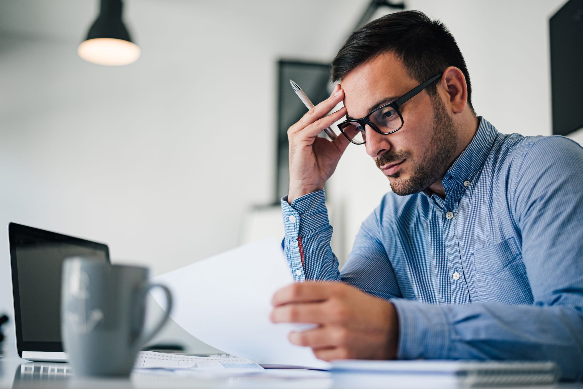 Man in glasses looking at papers, holding pen to head, laptop and mug on desk.