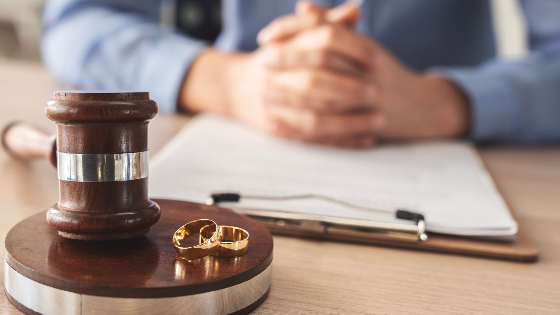 Gavel, wedding rings, and document on desk; person in background with clasped hands.