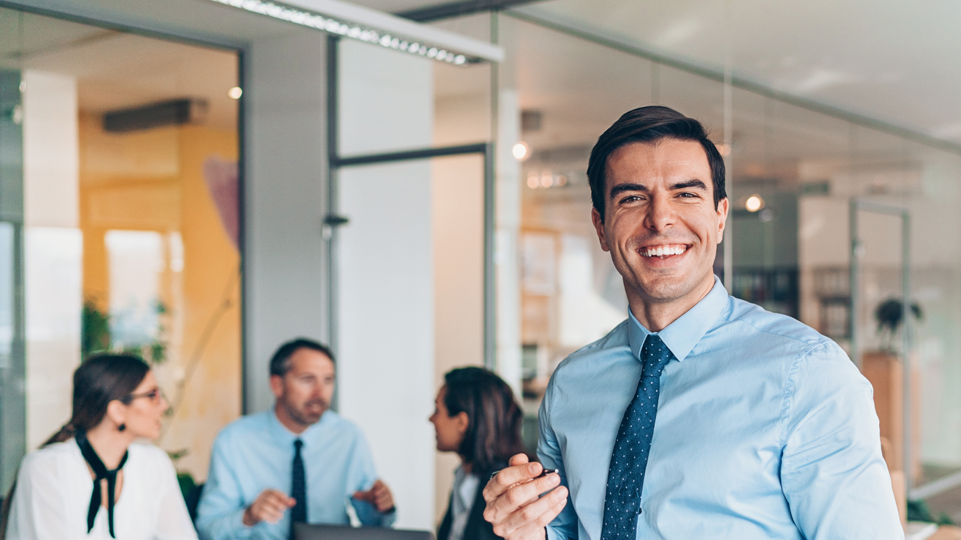 A man in a blue shirt and tie is smiling in front of a group of people in an office.