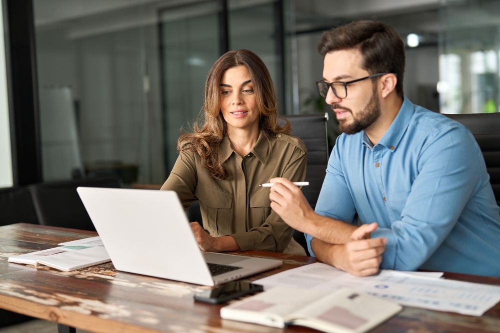 A man and a woman are sitting at a table looking at a laptop computer.