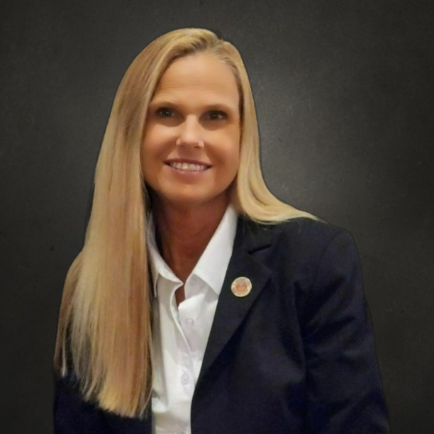 A woman in a black jacket and white shirt is smiling in front of a fireplace.