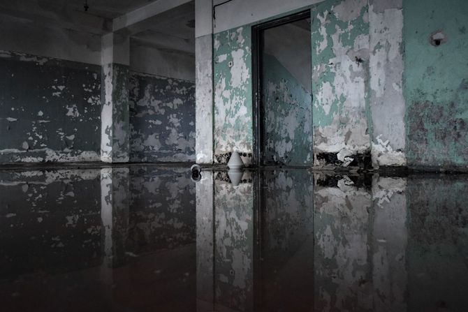 Dim, peeling-painted room with a reflective flooded floor and a closed door.