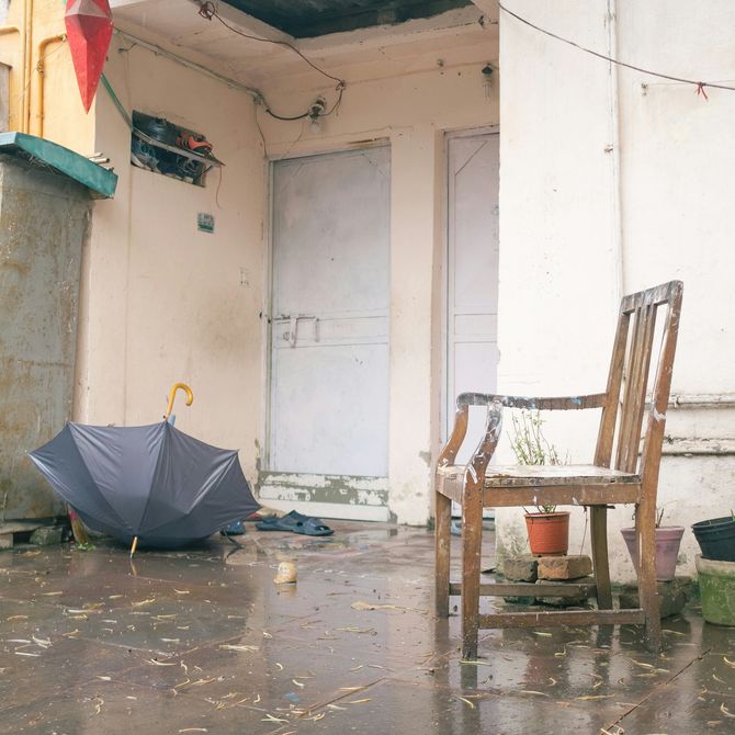 Rainy porch with a closed white door, a wooden chair, and an open umbrella on the wet floor.