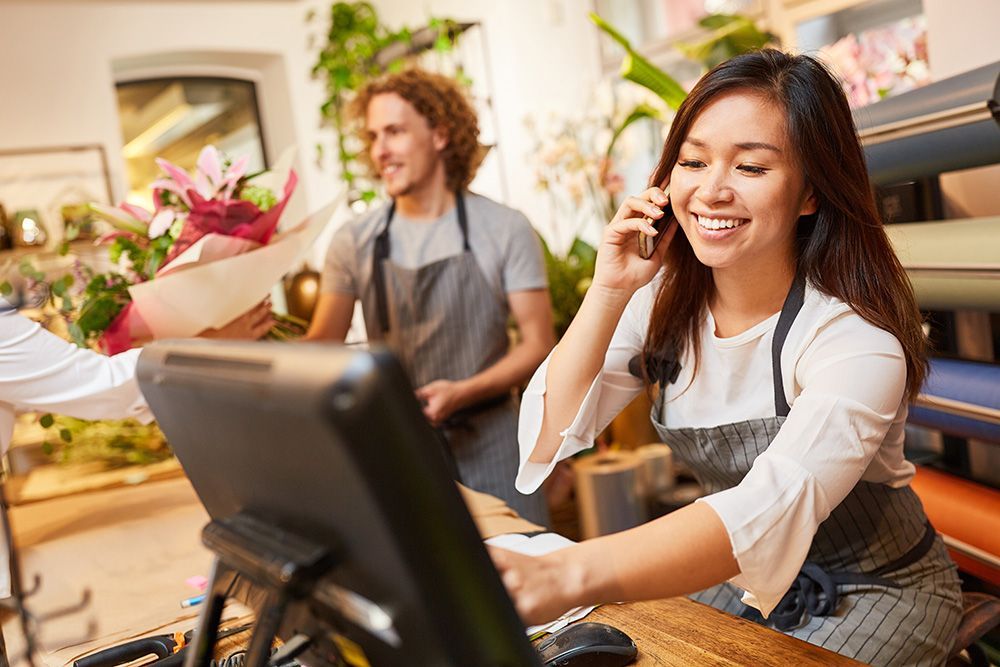 A woman is sitting at a counter in a flower shop talking on a cell phone.