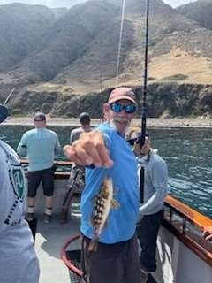Man on boat holding a small fish, smiling, near mountains, wearing a blue shirt and sunglasses.