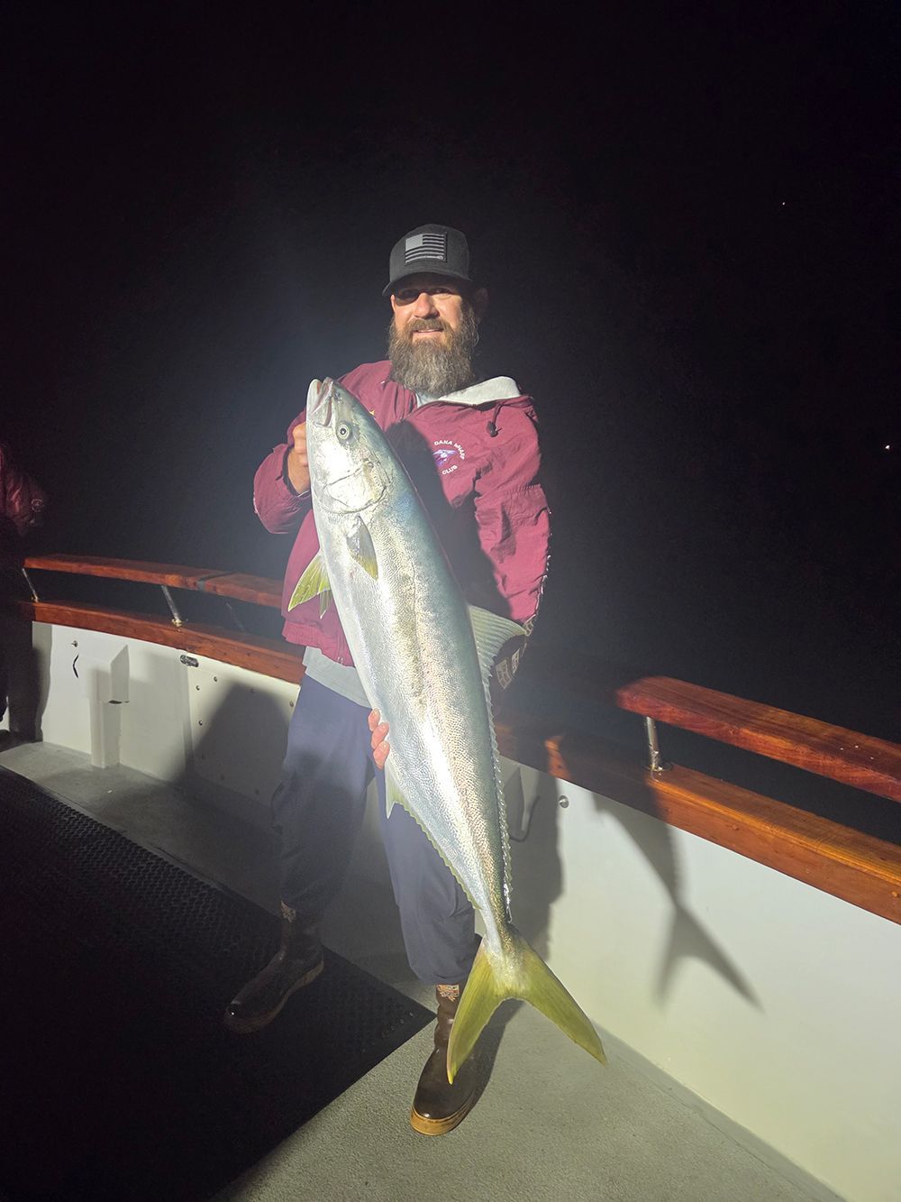 Man on a boat holding a large silver fish with yellow tail. Nighttime, wearing a cap, jacket.