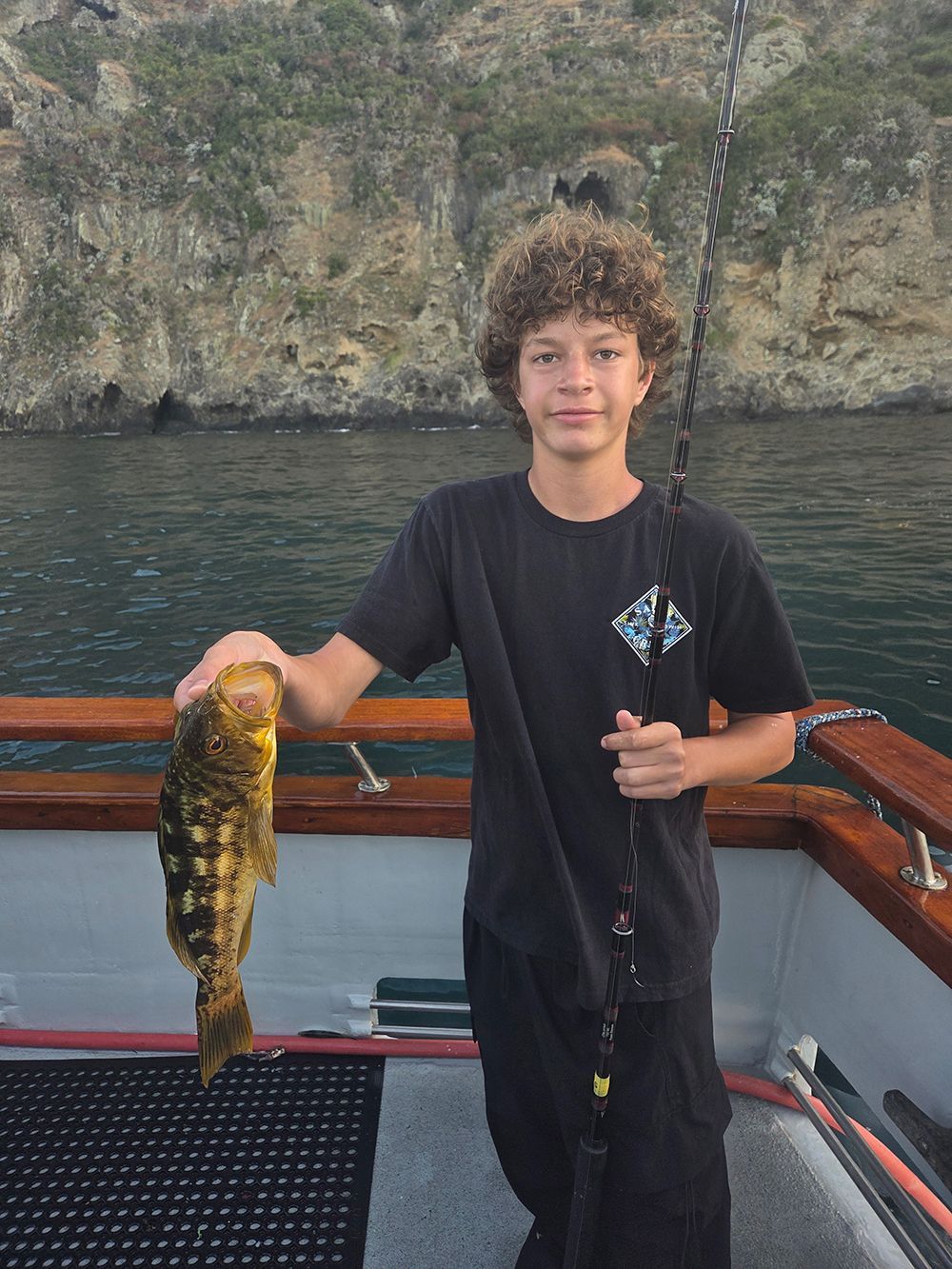 Young boy holding a yellow-brown fish on a boat, against a cliffside background.