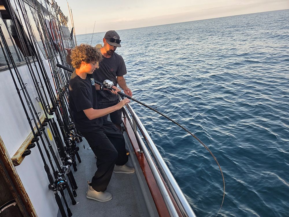 Boy fishing on a boat, assisted by an adult. Ocean in background, rods lined along the boat's side.