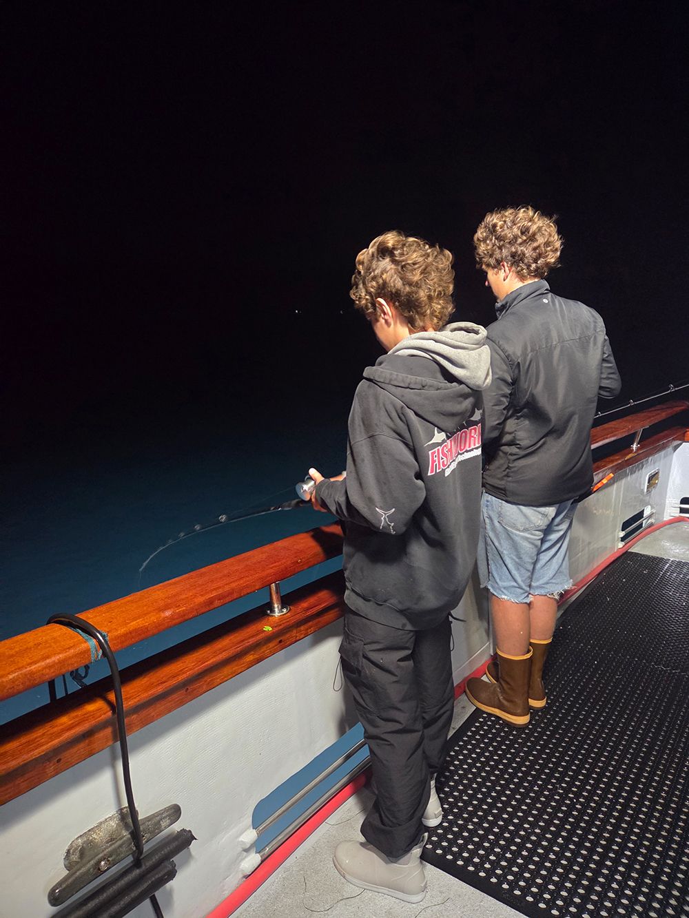 Two boys fishing at night from a boat. One boy wears black pants/jacket. The other wears shorts and boots.