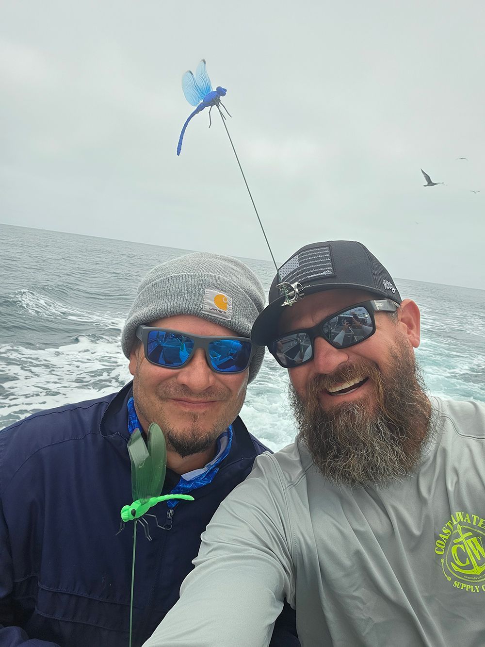 Two men smiling on a boat, fishing with blue dragonfly and green lure, overcast sky.
