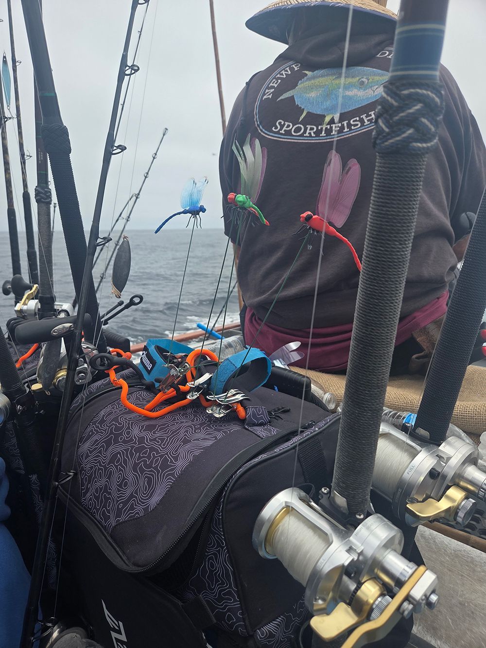 Fisherman on a boat with fishing rods and tackle bag, colorful lures hanging; overcast sky.