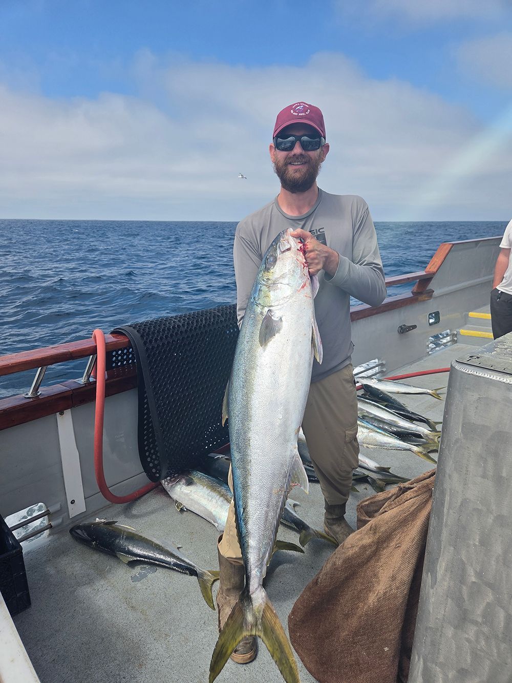 Man on boat holds up a large, silvery fish. Ocean in background.