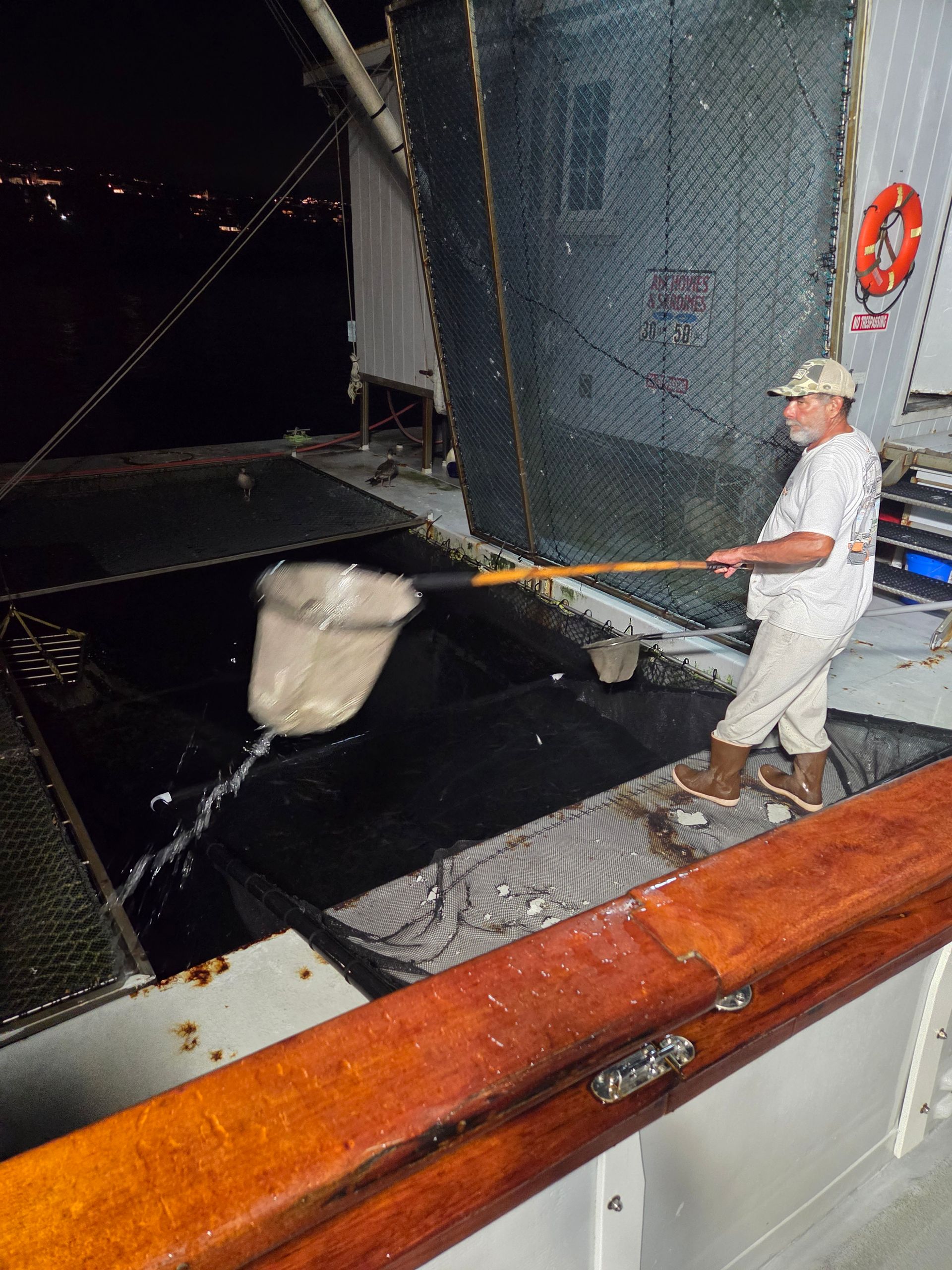 Man on a boat using a net to scoop up water. Dark setting, wooden railing, metal and netting visible.