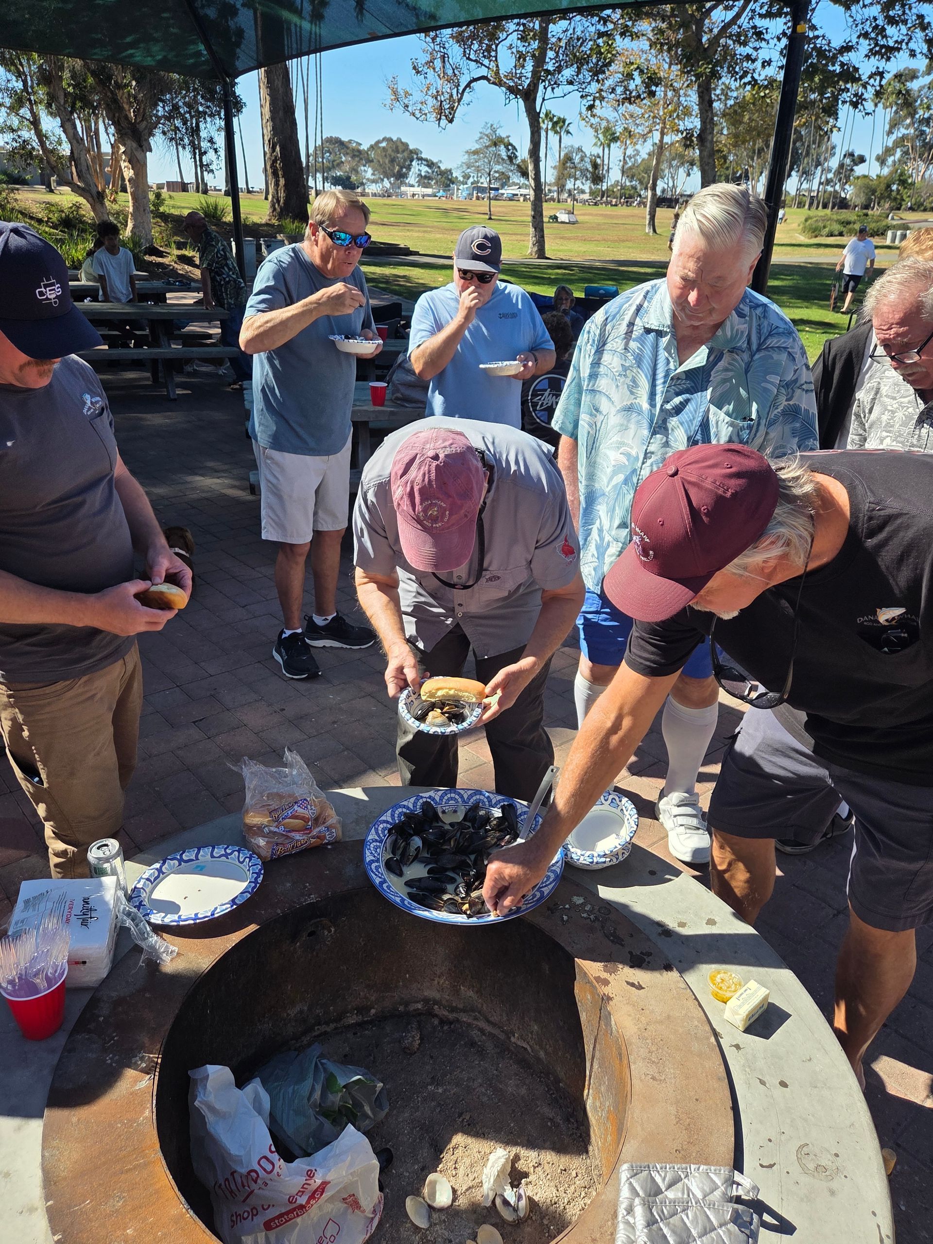 Men gather around a fire pit, serving food from plates in a park on a sunny day.