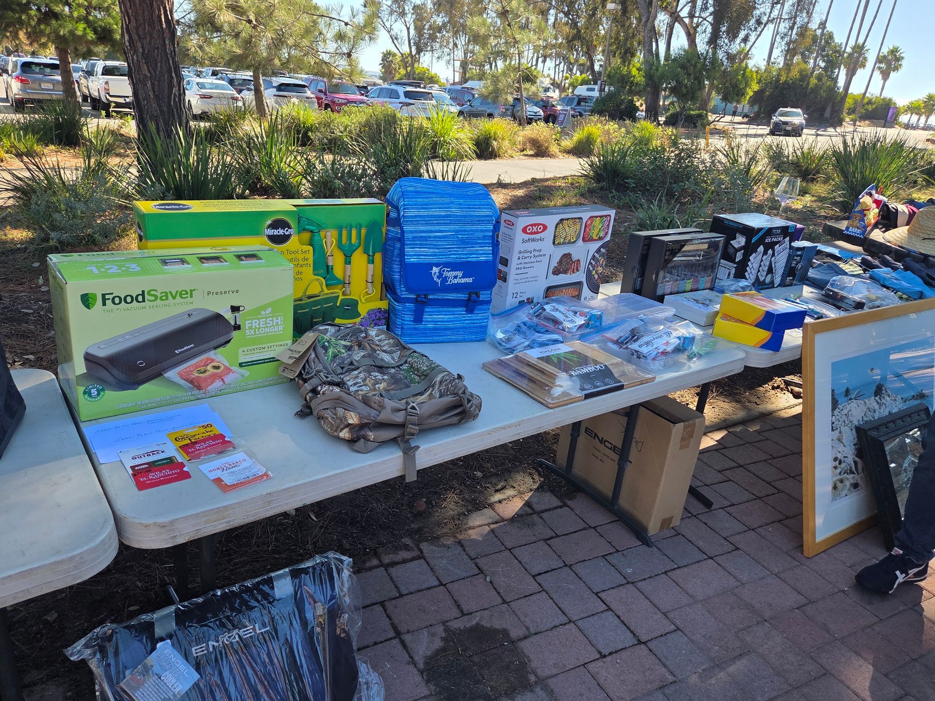 Garage sale with items on a table outside. Various boxes and bags with merchandise are displayed.