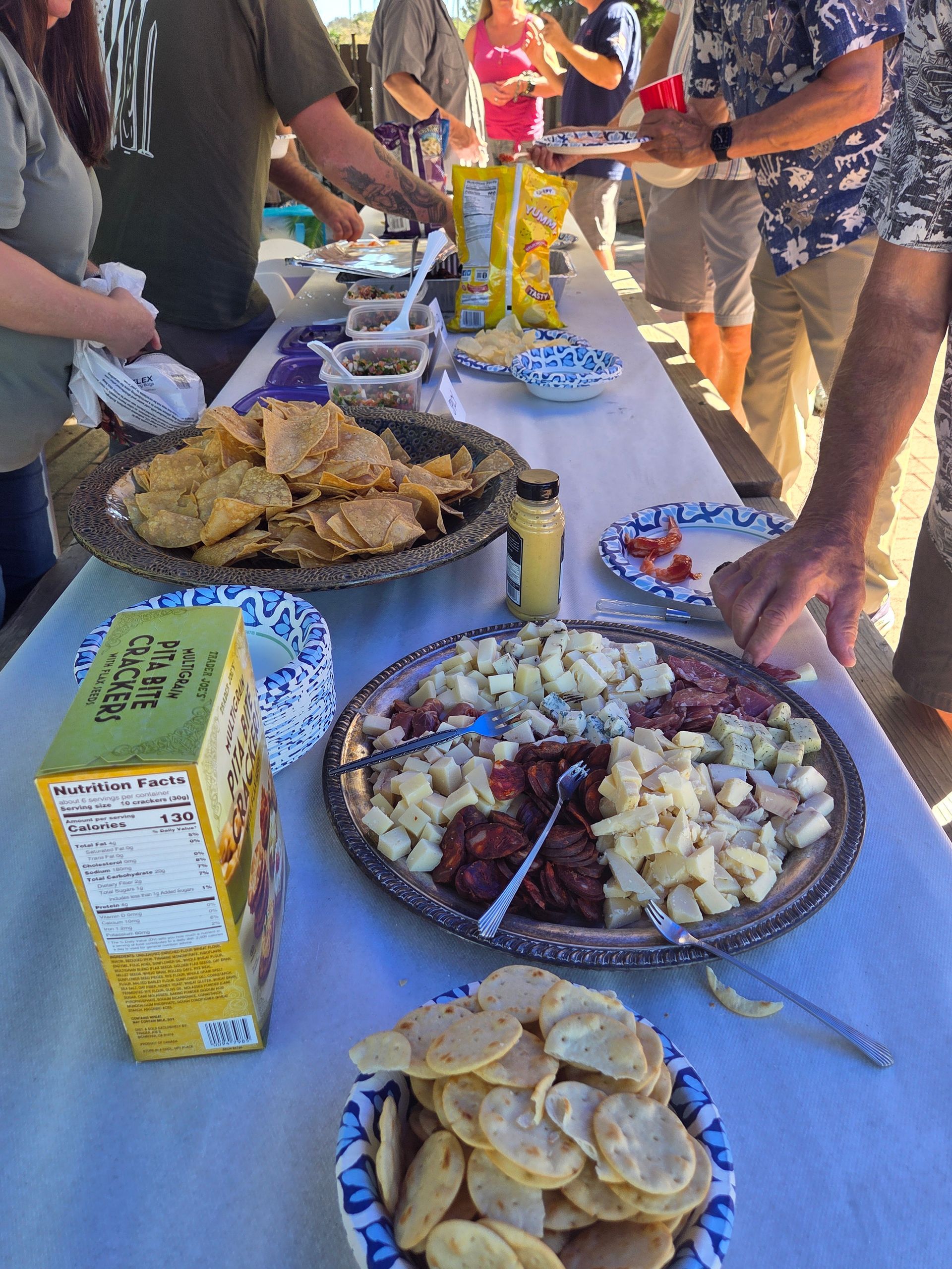 People at an outdoor food table with chips, dips, and other snacks on a white tablecloth.
