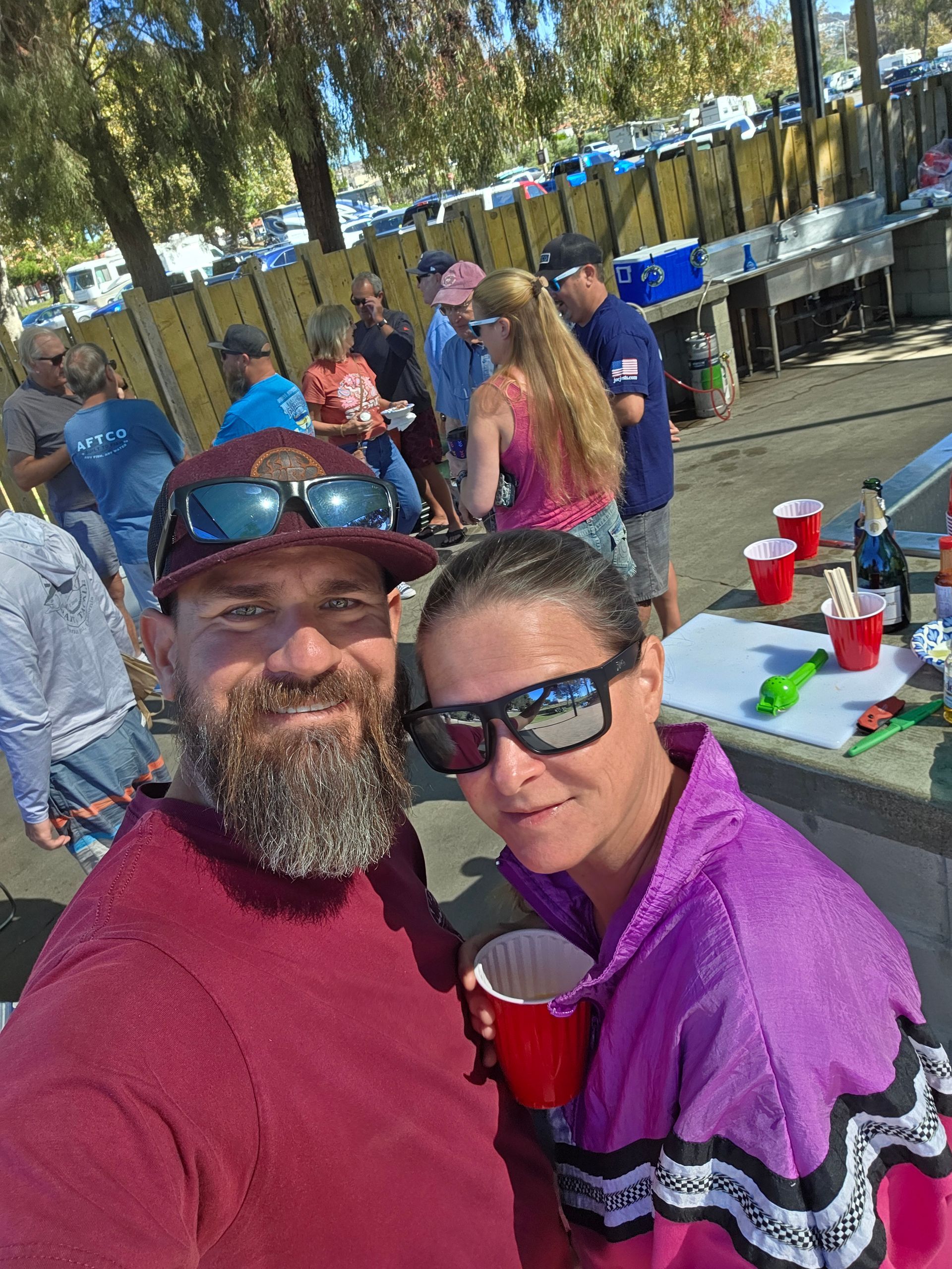 A man and woman with drinks pose for a selfie at an outdoor gathering. People are in the background.