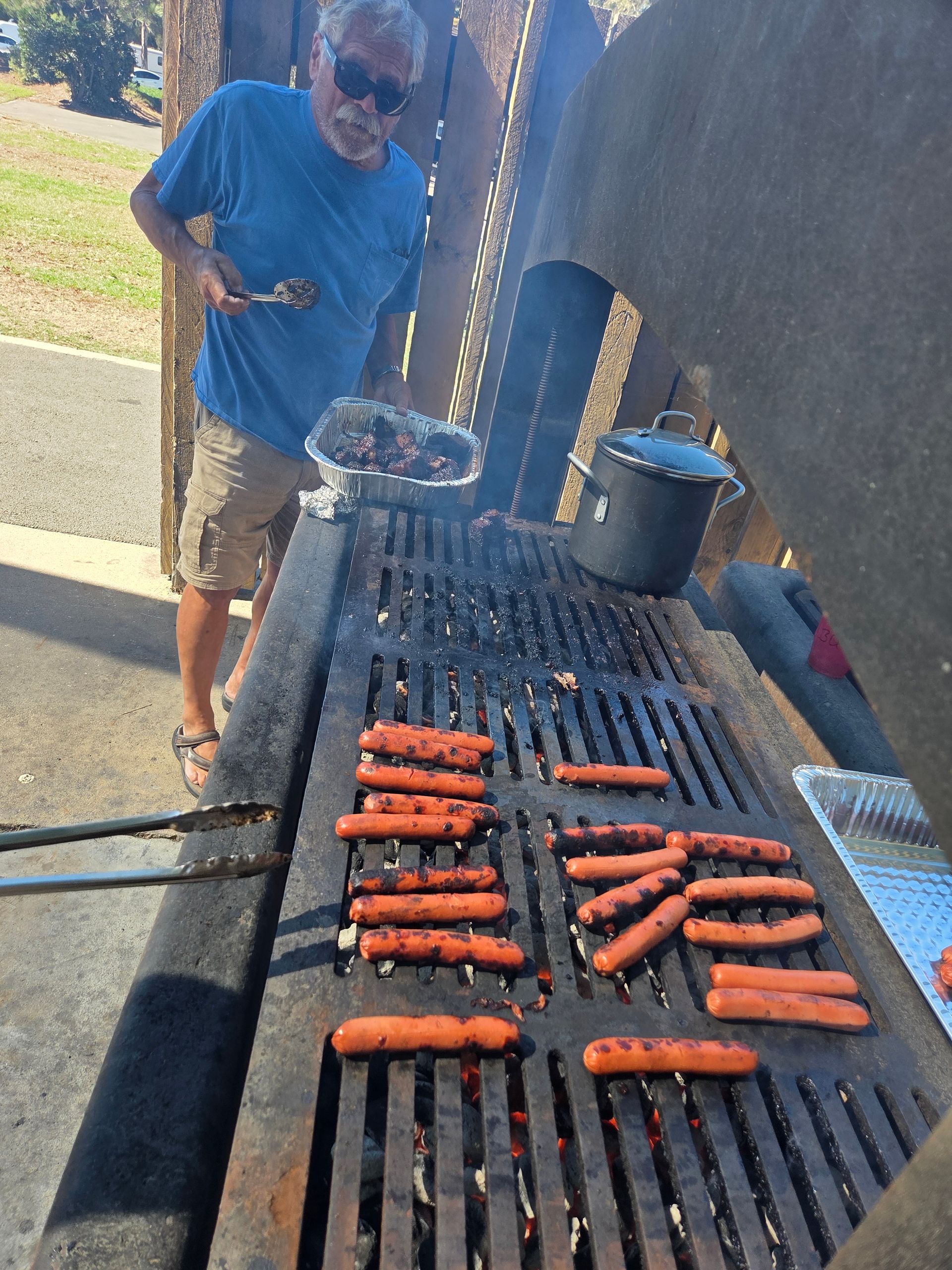 Man grilling hot dogs on a large outdoor grill, wearing sunglasses and a blue shirt, in a sunny setting.