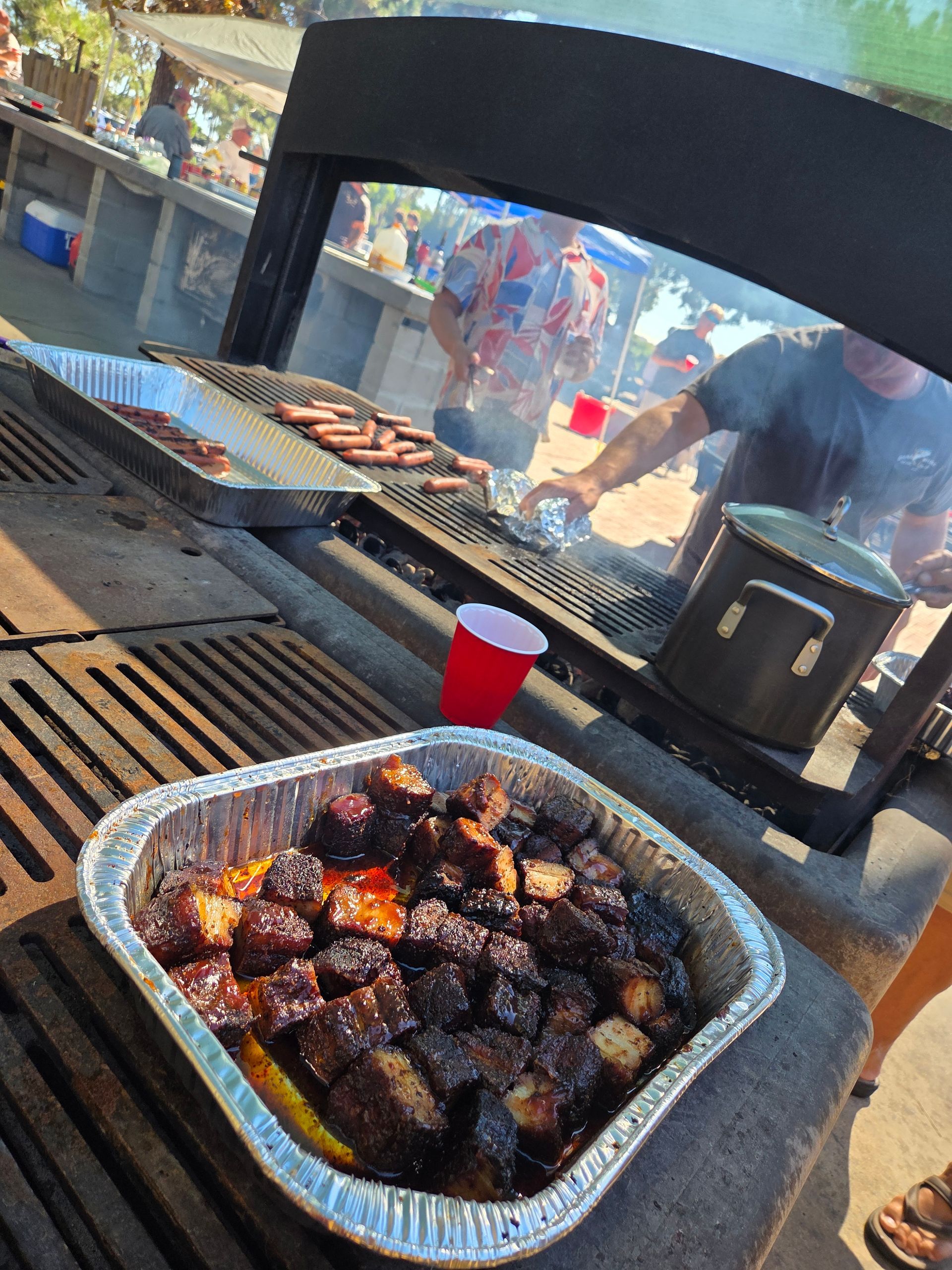 Smoked meat in foil pan on grill, people cooking. Outdoor event.