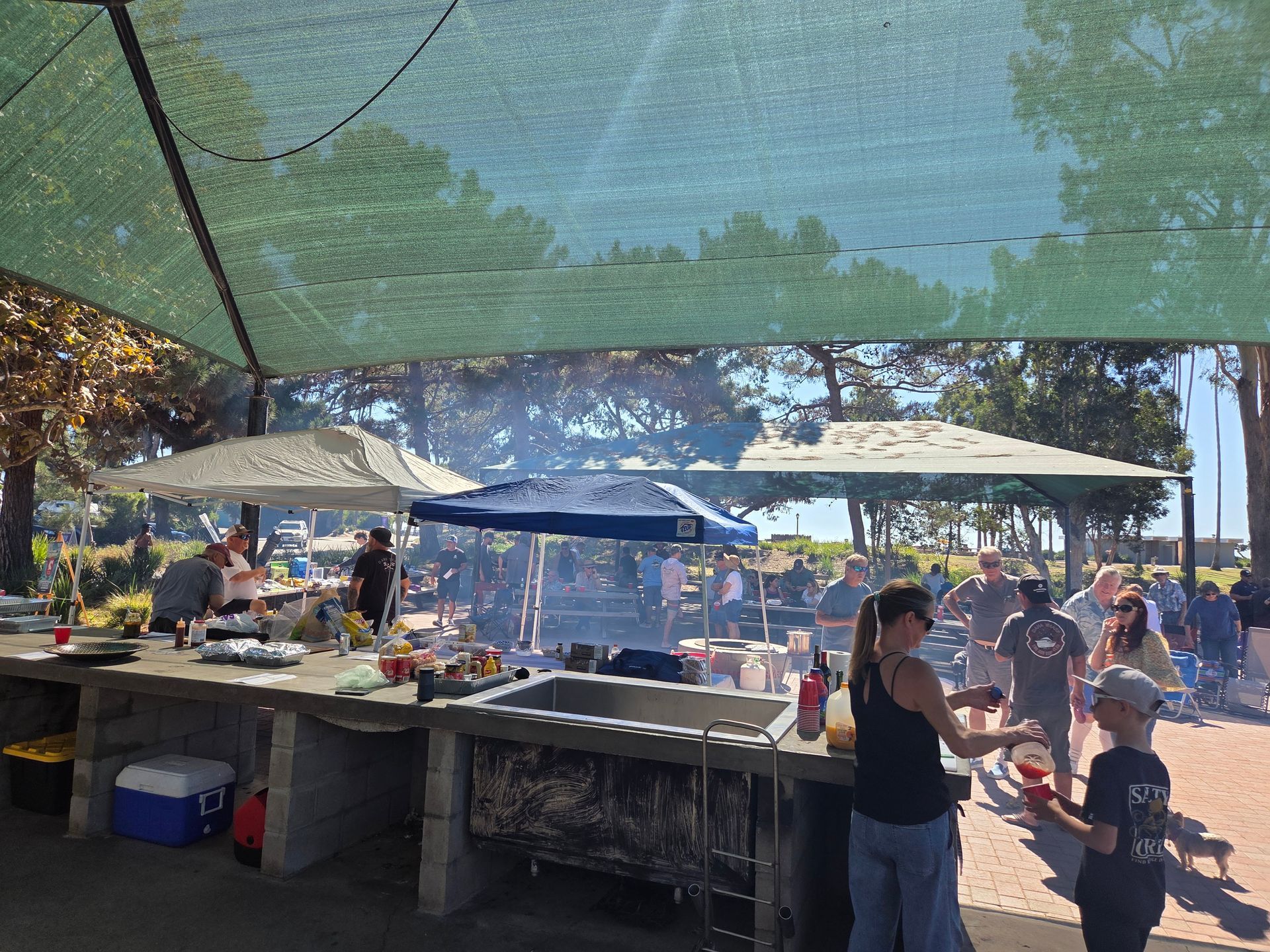 Outdoor gathering under a shade structure with people preparing food, and smoke rising.