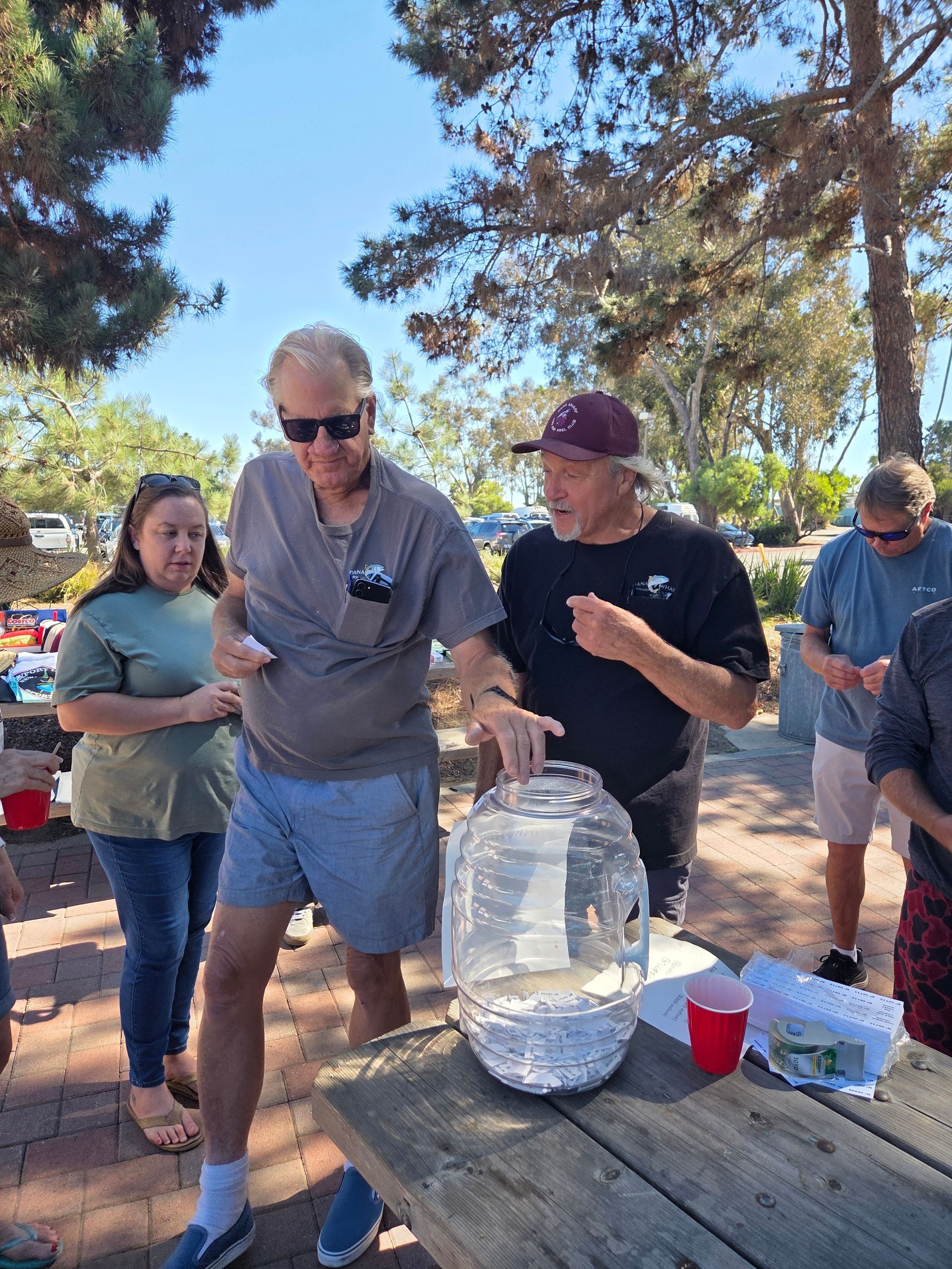 People by a picnic table, man pouring water from a large jug, sunny outdoor setting.