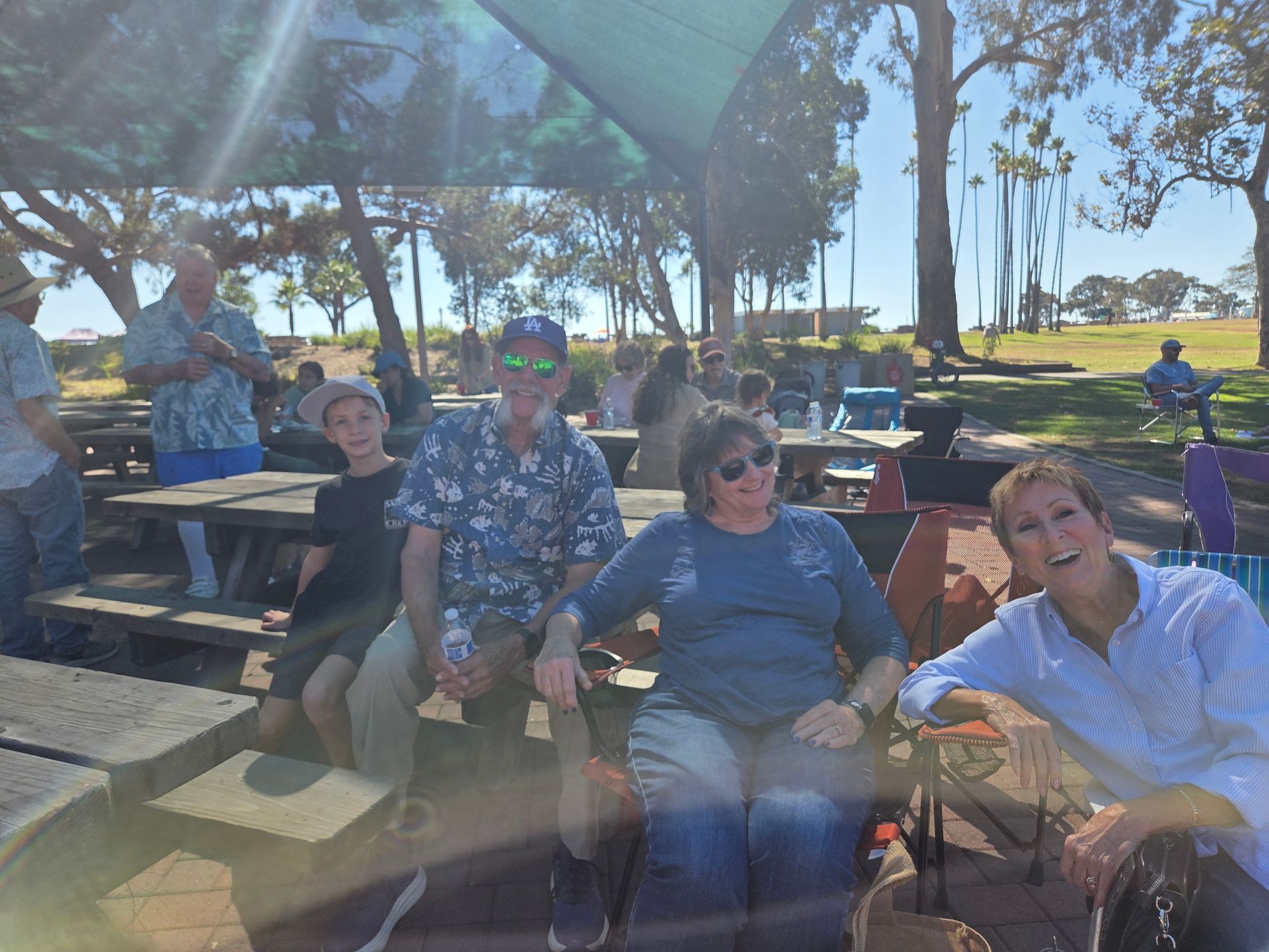 Group of people sitting at picnic tables in a park, smiling. Blue sky, trees, and sun overhead.