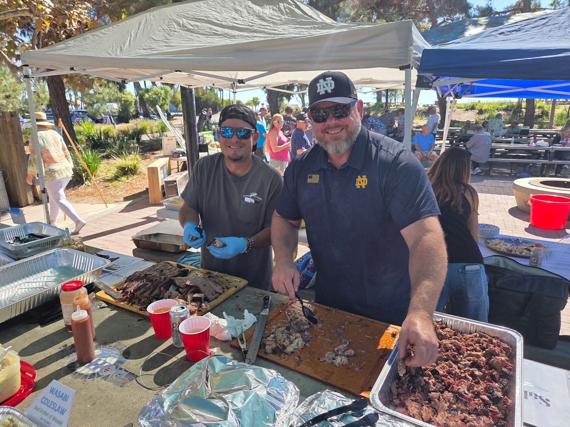 Two people serving food from a barbecue stand outdoors; meats on display, sunny day.