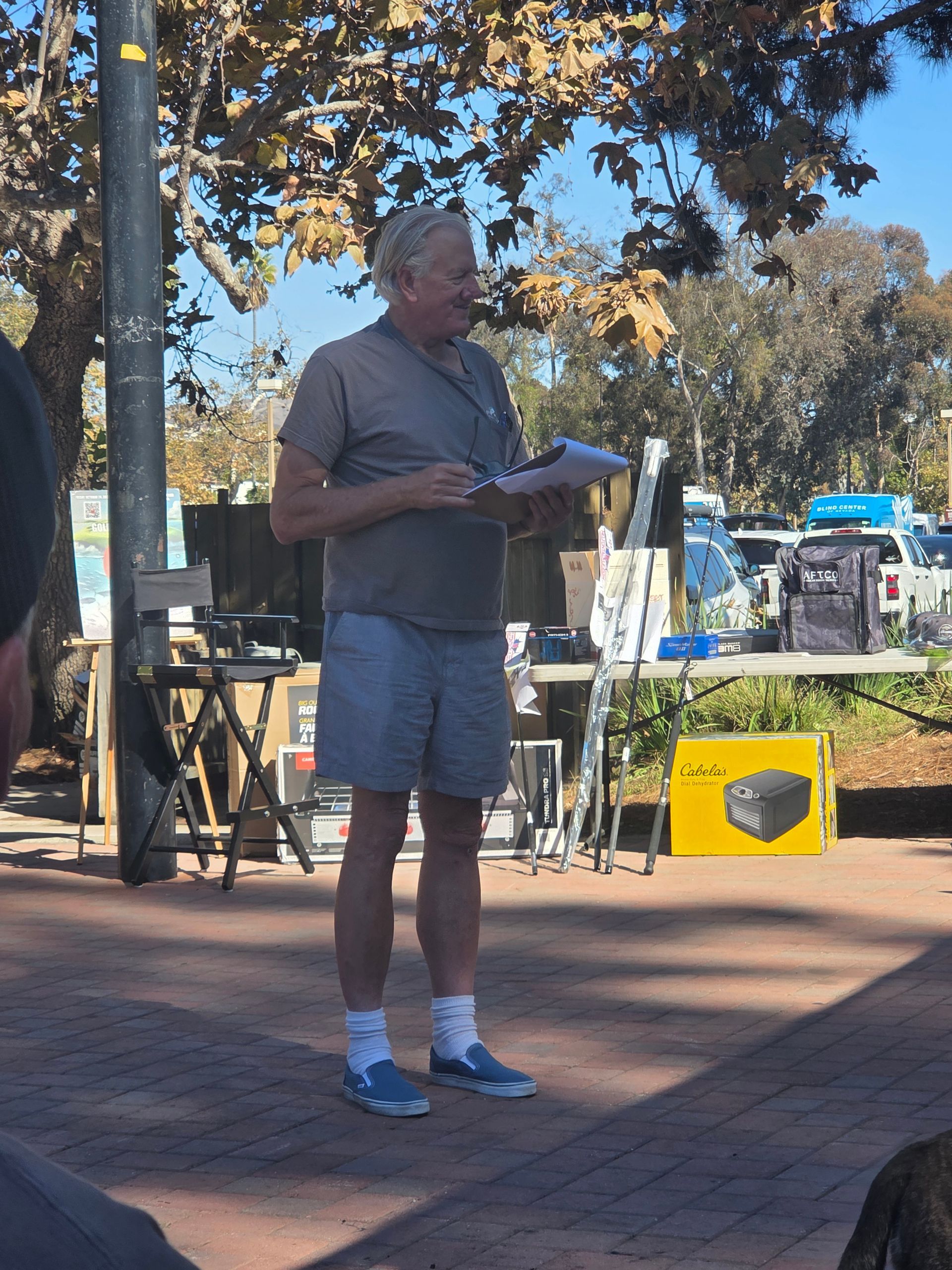Man in shorts speaks into an outdoor crowd, holding papers. Tables, products, and a sunny background.