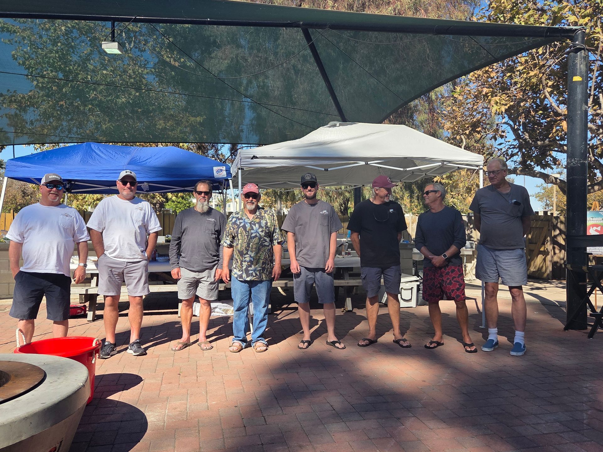 Group of eight men standing under shade structures outdoors.