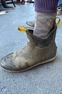 Dirty green rubber boot with sock pulled up; yellow pull tabs.