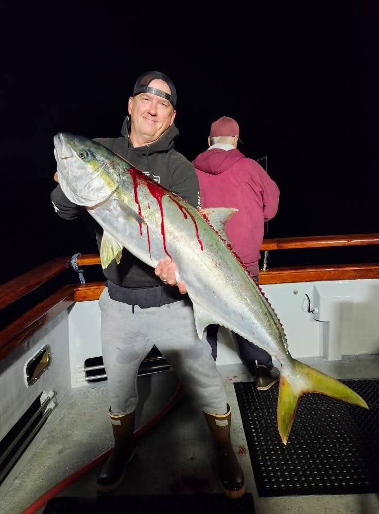 Man on boat holding large yellowtail fish, covered in blood. Nighttime setting.