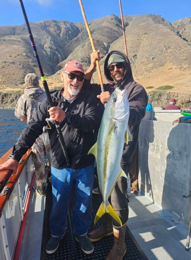 Two men on a boat hold up a large yellowtail fish, mountains in the background.