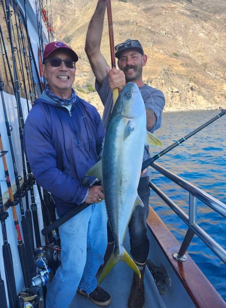 Two men on a boat display a large yellowtail fish they caught, with a mountain backdrop.