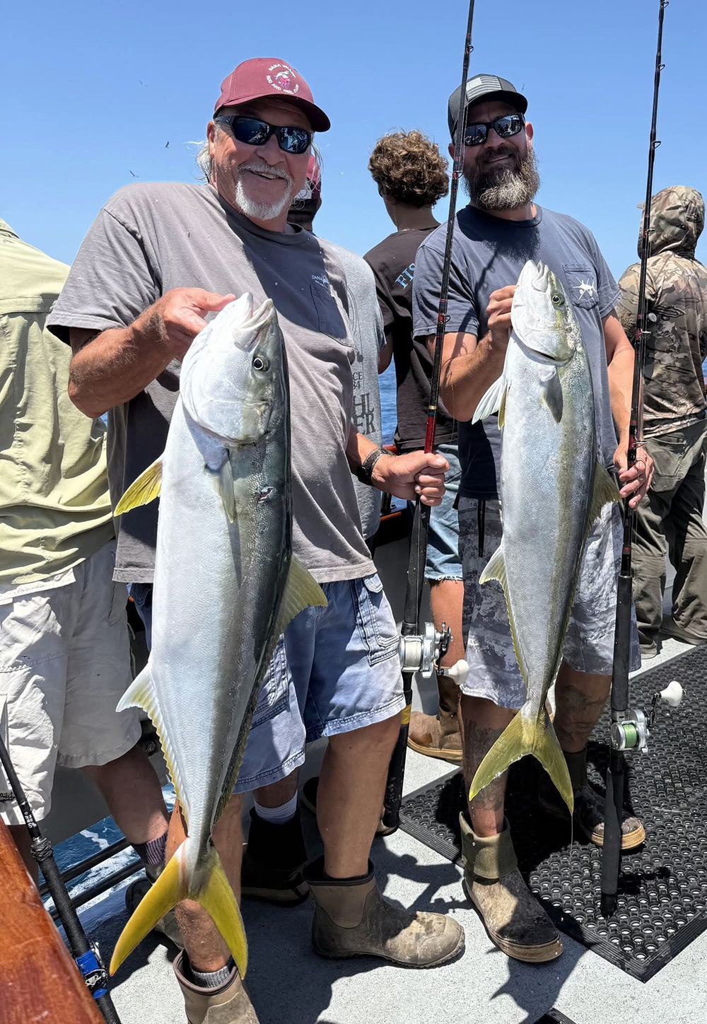 Two men on a boat, holding up large yellowtail fish they caught. Bright sun, blue water.