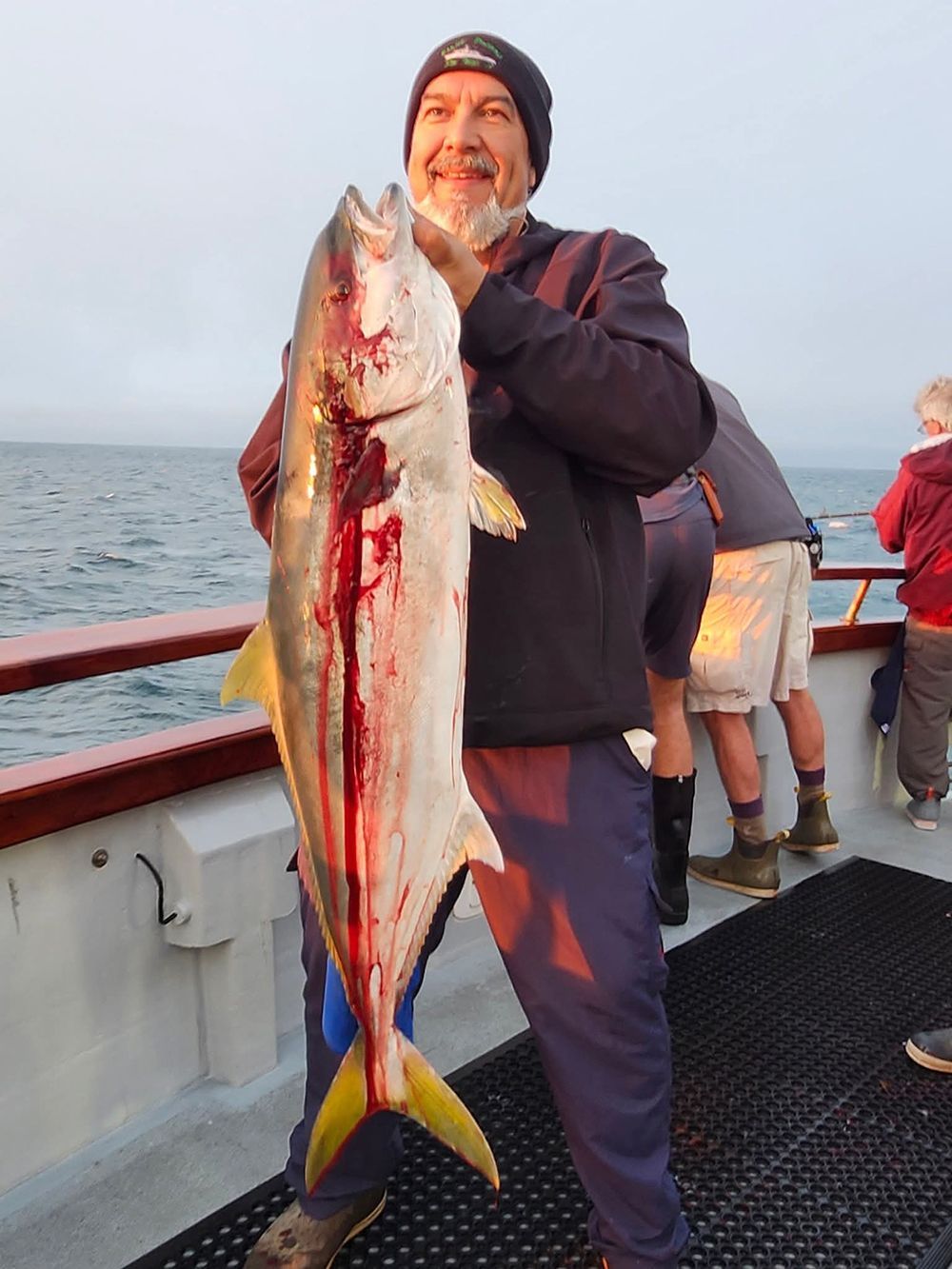 Man on boat holds up a large yellowtail fish, covered in blood.