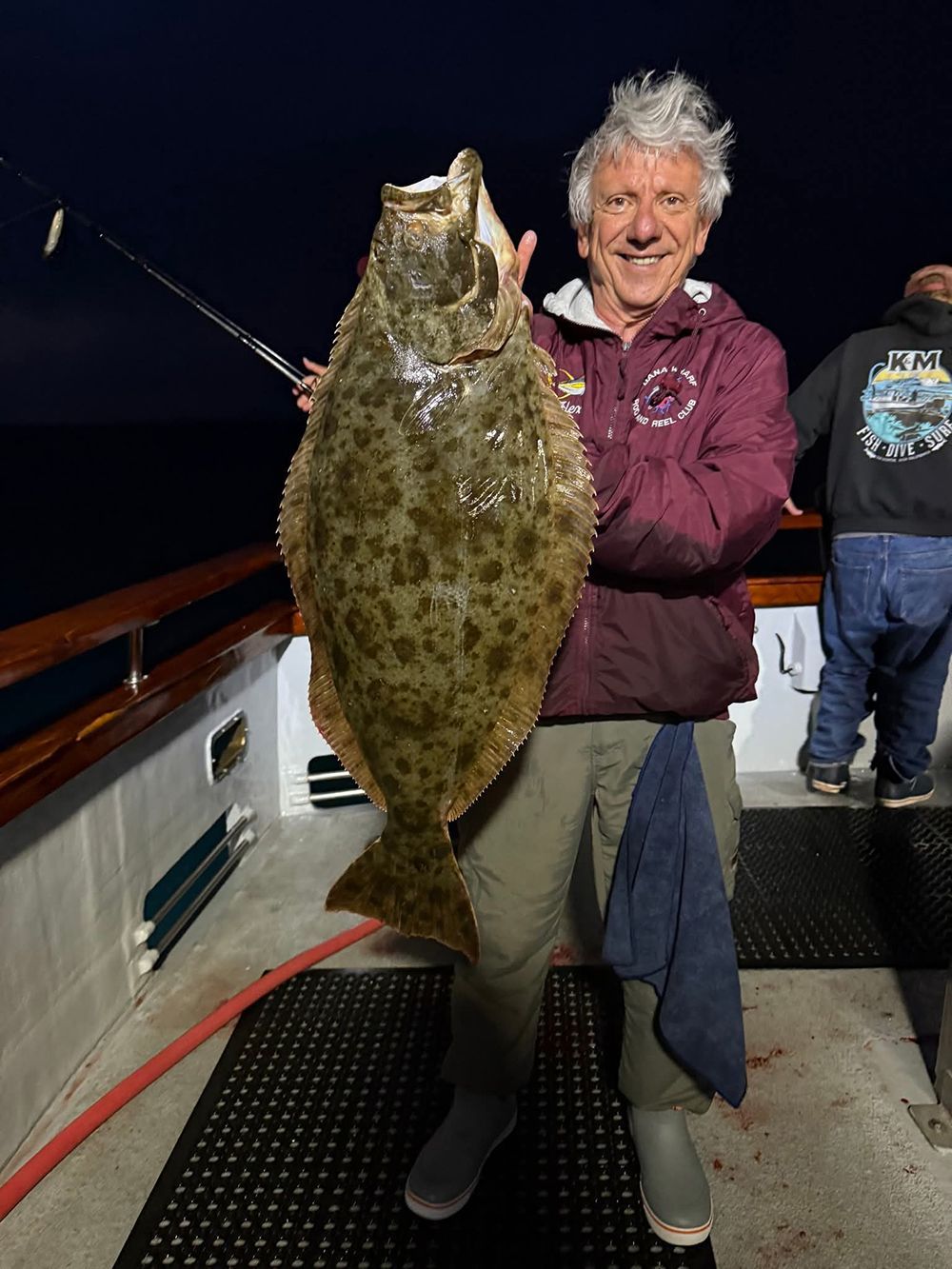 Man on a boat holds a large halibut, smiling. He wears a maroon jacket and khaki pants.