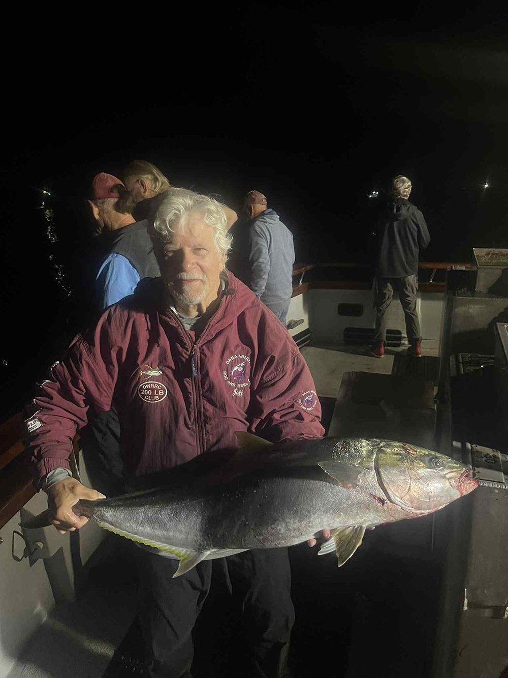 Man on a boat at night holds up a large yellowtail fish. Other people are visible on the boat.