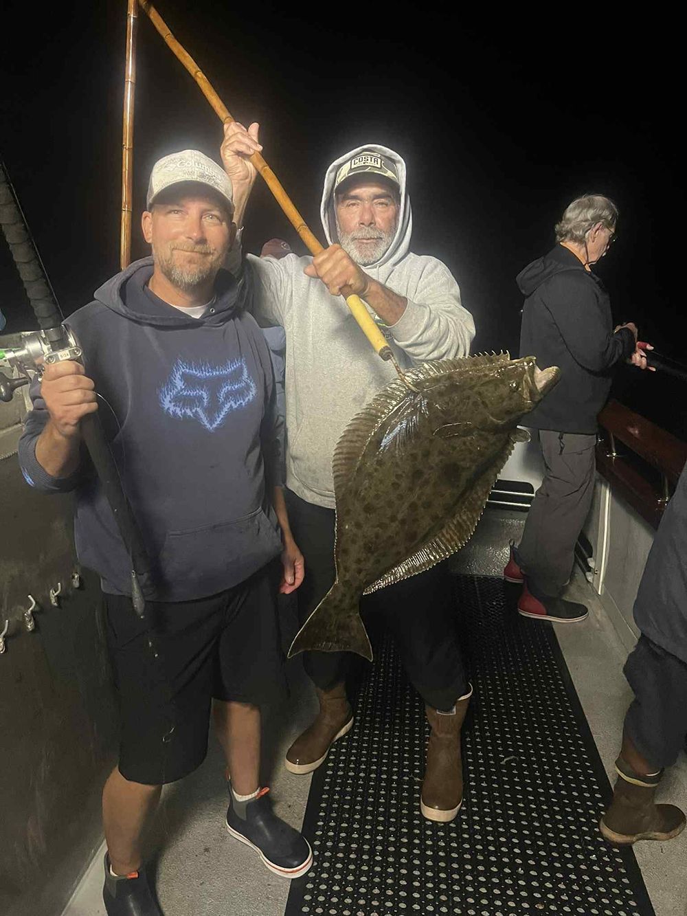 Two men on a boat holding a large speckled fish at night.