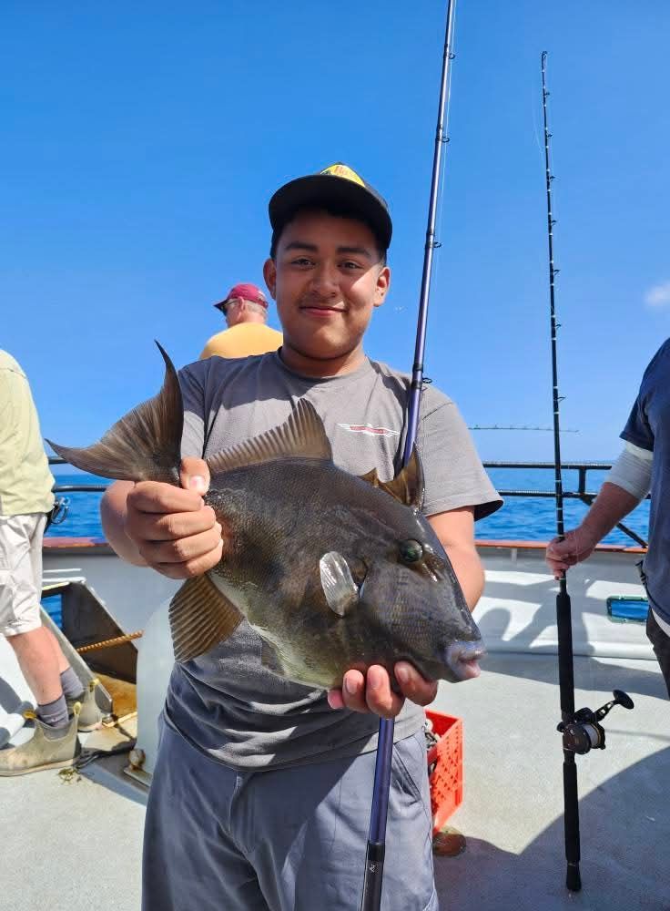 Young man smiling, holding a large dark triggerfish on a boat with fishing rods; ocean background.