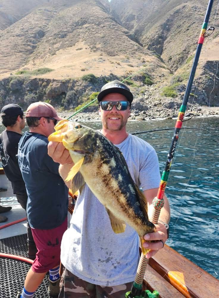 Man on boat smiles holding a yellow and brown fish, rod in hand, mountains in background.