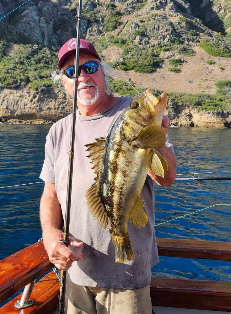 Man on boat holding large fish, smiling. Ocean setting, other people visible.