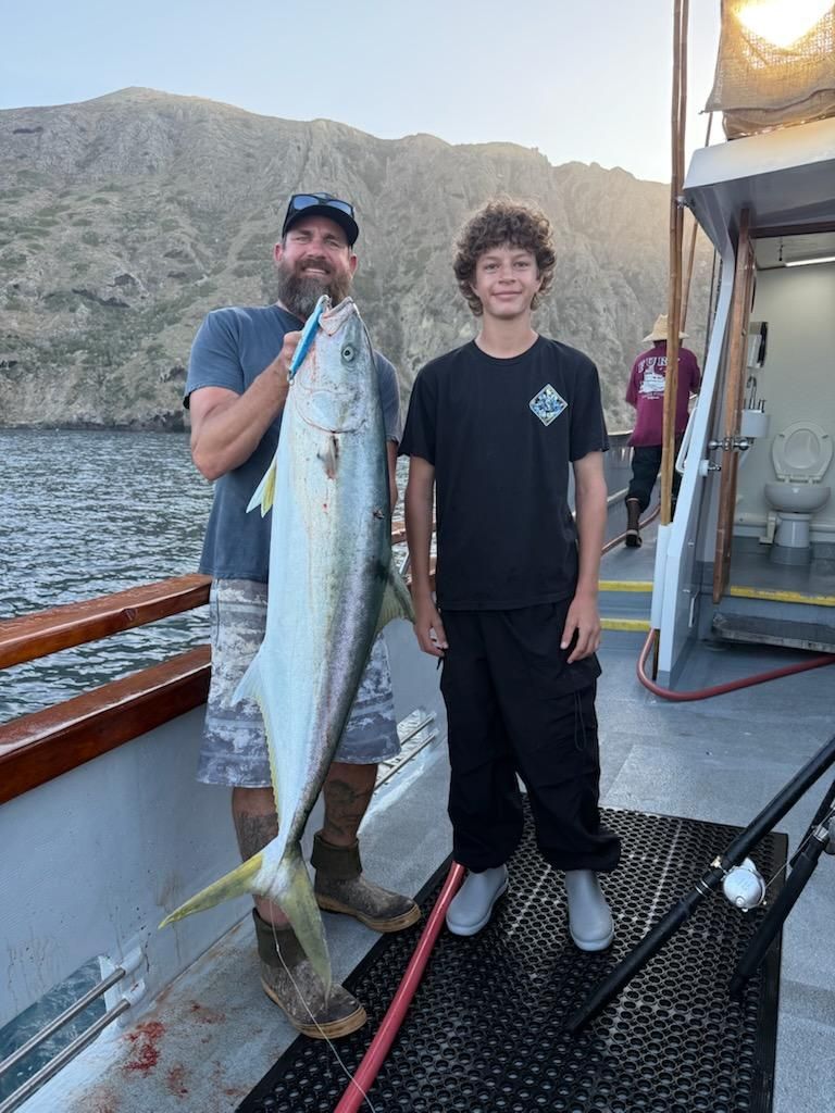 Man and boy on a boat holding a large yellowtail fish near mountains.