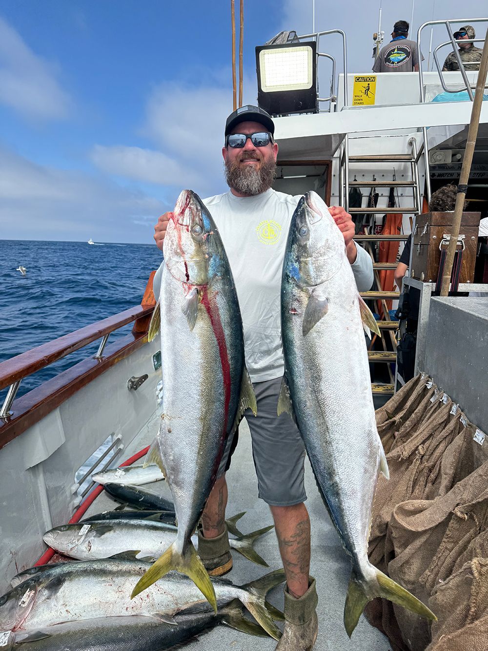 Man on boat holds up two large yellowtail fish, smiling. Other fish on deck.