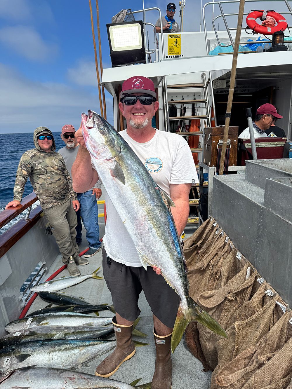 Man on a boat holds a large fish. Other anglers and fish are visible on the deck under a blue sky.