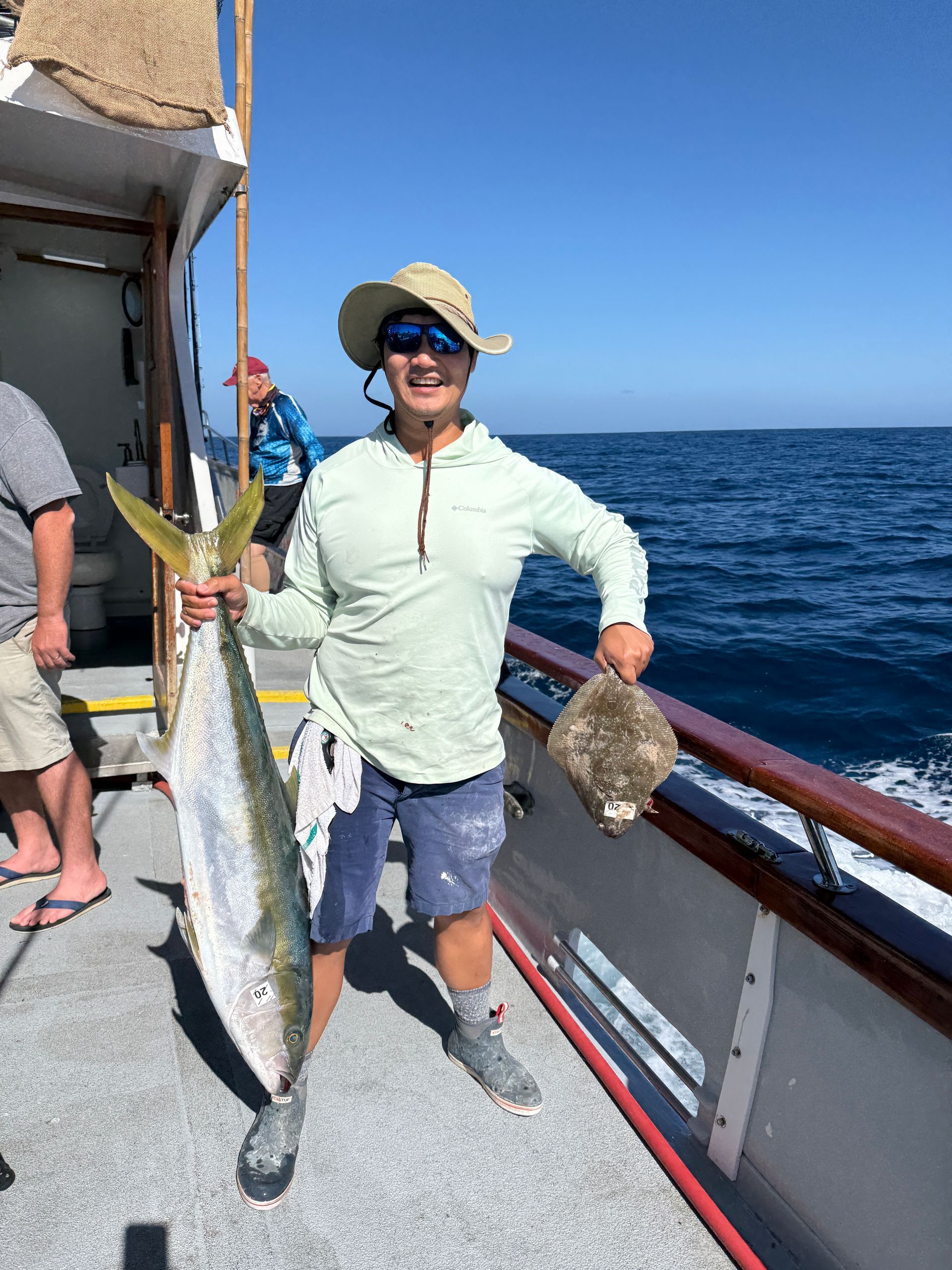 Man on a boat holds a large yellowtail fish and a smaller flatfish, ocean background.